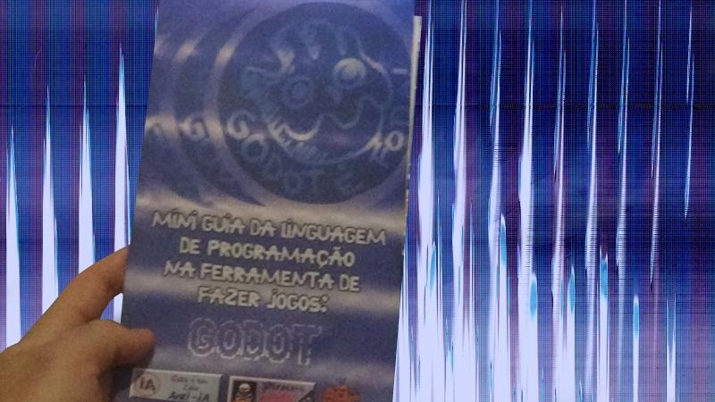 Foto editada de pessoa segurando um zine cujo sua capa tem uma ilustração da logo da Godot que é um robozinho com cabeça de engrenagem, a ilustração está dentro de um círculo escrito embaixo -Godot é foda!, este círculo parece estar se deslocando criando mais 3 cópias de si mesmo ao mesmo tempo que some. Abaixo disto está o título da zine: - Mini guia da linguagem de programação na ferramenta de fazer jogos: Godot. Abaixo do título tem a pixel art de 3 topeiras punks do solo escrito punkytrio, e dois botões escritos - Pirateie Agora! Free, - Este é um zine Anti-IA.

A mão segurando o zine está sem fundo, possuindo um fundo psicodélico de cores azul, roxo e branco.